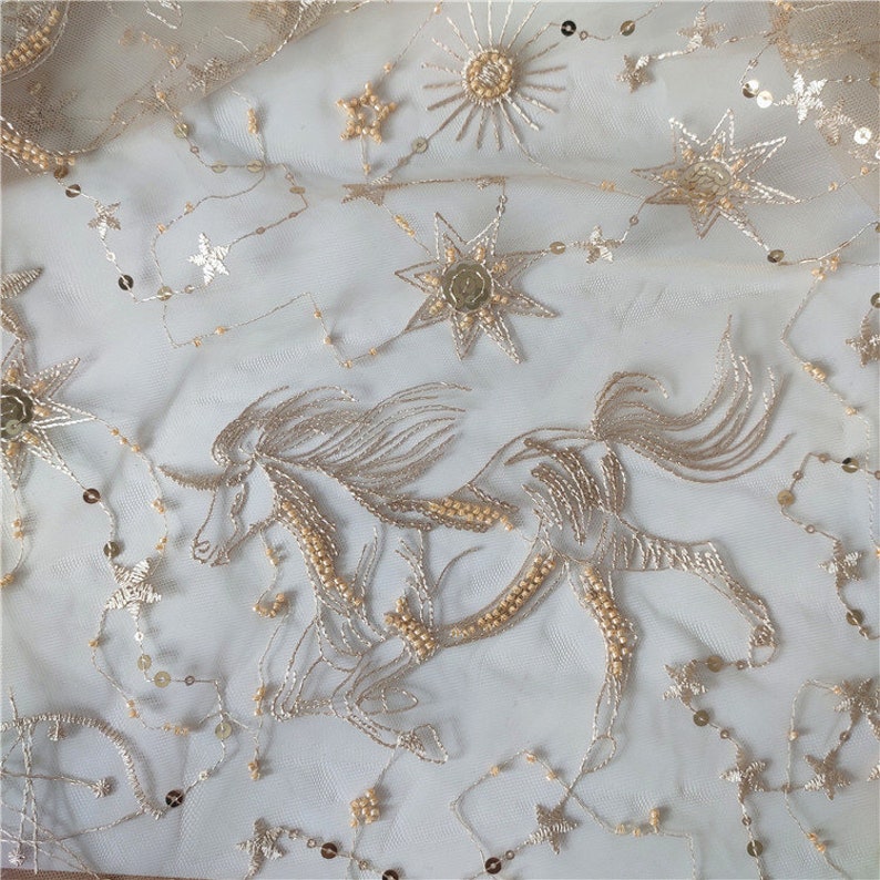 May include: Gold embroidered tulle fabric with a unicorn and stars design. The fabric is sheer and has a delicate texture.