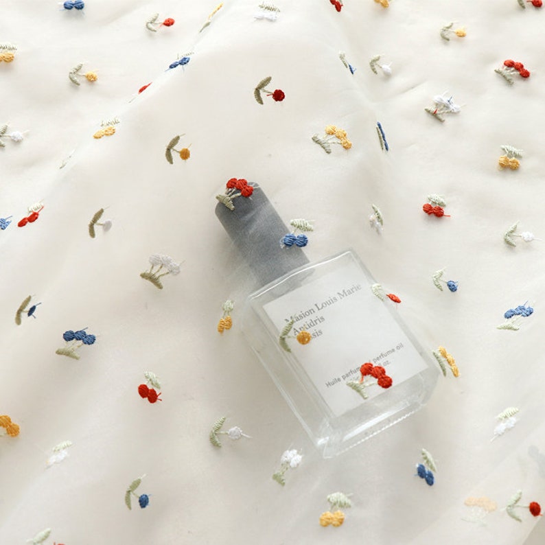 K&ouml;nnte beinhalten: Wei&szlig;er, durchsichtiger Stoff mit gestickten roten, gelben und blauen Kirschen. Der Stoff liegt auf einem Tisch mit einer durchsichtigen Glasflasche Parf&uuml;m mit dem Text "Maison Louis Marie" und "Anisdris" auf dem Etikett.