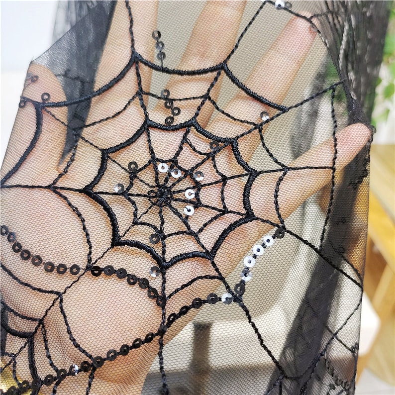 May include: A sheer black mesh fabric featuring an embroidered spider web pattern. The design is accented with small, circular black sequins stitched along the web lines, creating a textured and reflective surface on the translucent material.