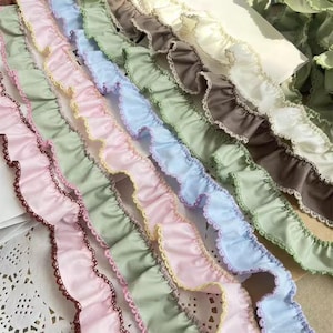 Puede incluir: Primer plano de filas de adornos de tela con volantes en varios colores pastel. Los colores incluyen rosa, azul, verde y crema. Cada tira tiene un borde contrastante, algunas con ribete de encaje. La tela parece suave y ligera.