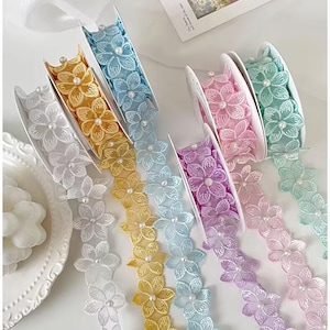 May include: A collection of floral lace ribbons in various pastel colors, including white, yellow, blue, pink, purple, and green. Each ribbon features a repeating flower design with a pearl accent in the center of each flower. The ribbons are displayed on white spools.