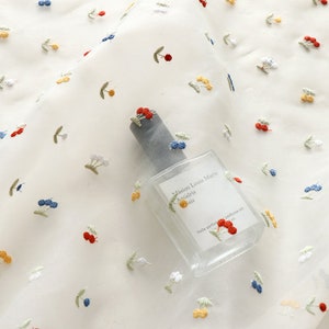 K&ouml;nnte beinhalten: Wei&szlig;er, durchsichtiger Stoff mit gestickten roten, gelben und blauen Kirschen. Eine durchsichtige Glasflasche mit dem Text "Maison Louis Marie" und "Anisdris" liegt auf dem Stoff.