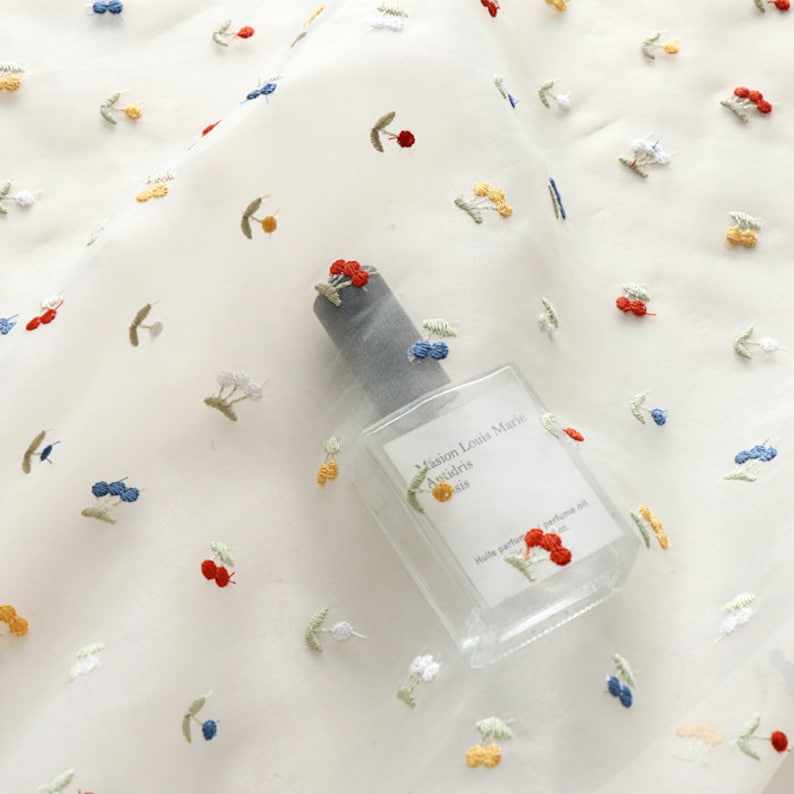 K&ouml;nnte beinhalten: Wei&szlig;er, durchsichtiger Stoff mit gestickten roten, gelben und blauen Kirschen. Eine durchsichtige Glasflasche mit dem Text "Maison Louis Marie" und "Anisdris" liegt auf dem Stoff.
