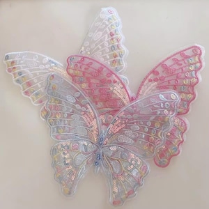 Puede incluir: Primer plano de varios apliques con forma de mariposa. Los apliques están hechos de tela transparente y decorados con lentejuelas y bordados. Los colores incluyen blanco, rosa y azul con acentos iridiscentes. Las alas están detalladas con patrones intrincados.