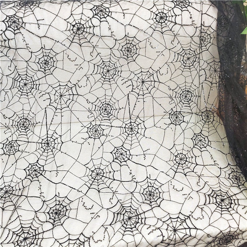 May include: A black spiderweb patterned fabric featuring intricate web designs with small, reflective black sequins stitched along the lines. The sheer, lightweight material displays a repeating pattern of interconnected webs across the surface.