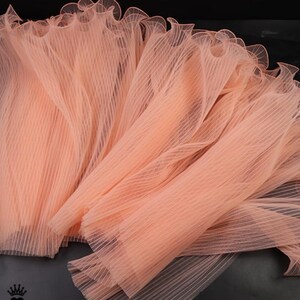Peut inclure: Gros plan sur plusieurs morceaux de tissu pliss&eacute; de couleur p&ecirc;che. Le tissu a une qualit&eacute; d&eacute;licate et transparente avec un bord &agrave; volants. Le mat&eacute;riau est dispos&eacute; en couches, cr&eacute;ant une sensation de profondeur et de texture. Id&eacute;al pour les projets d'artisanat ou de mode.