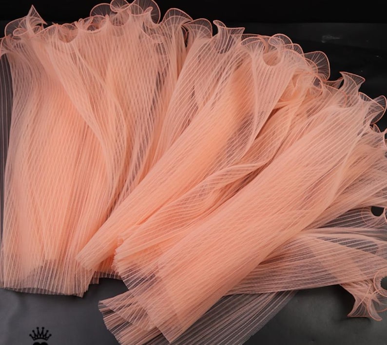 Peut inclure: Gros plan sur plusieurs morceaux de tissu pliss&eacute; de couleur p&ecirc;che. Le tissu a une qualit&eacute; d&eacute;licate et transparente avec un bord &agrave; volants. Le mat&eacute;riau est dispos&eacute; en couches, cr&eacute;ant une sensation de profondeur et de texture. Id&eacute;al pour les projets d'artisanat ou de mode.