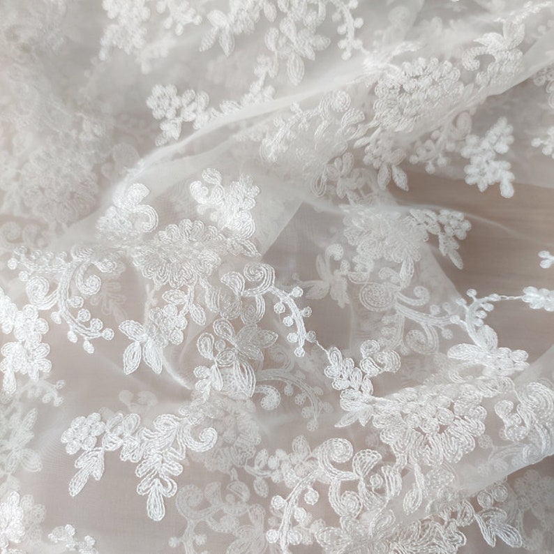 May include: White floral lace fabric with a delicate pattern. The lace is sheer and has a soft, flowing texture.