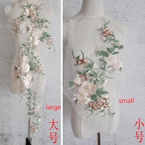 Puede incluir: Dos apliques florales bordados, uno grande y otro pequeño, con rosas blancas y rosas con hojas verdes. Los apliques se muestran sobre un fondo de tela de malla blanca.
