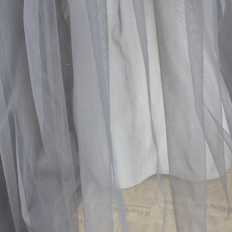 Pu&ograve; includere: Primo piano di un tessuto di tulle grigio trasparente. Il tessuto &egrave; morbido e delicato, con una superficie leggermente strutturata.