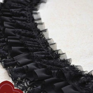 Könnte beinhalten: Schwarze Spitzenborte mit einem gekräuselten Rand. Die Borte ist aus einem weichen, durchsichtigen Stoff gefertigt und hat einen zarten, femininen Look.