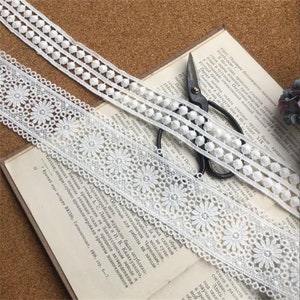 May include: Two white lace trims, one with a scalloped edge and the other with a row of white pom-poms. The trims are laid out on a book with a pair of scissors.