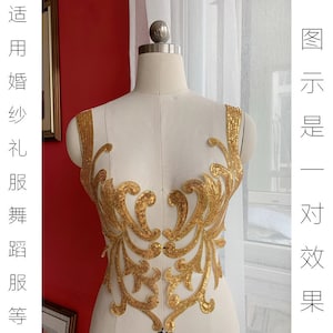 May include: A pair of gold sequined appliques, displayed on a mannequin. The appliques feature an ornate, symmetrical design with swirling patterns. The text on the left side of the image is in Chinese. The appliques are designed for bridal wear and dance costumes.