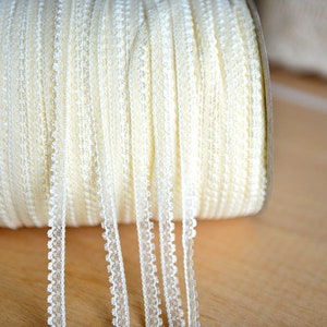 Könnte beinhalten: Eine Spule mit weißer Spitzenborte mit einem zarten gewellten Rand. Die Spitze ist aufgerollt und die Enden hängen herunter.
