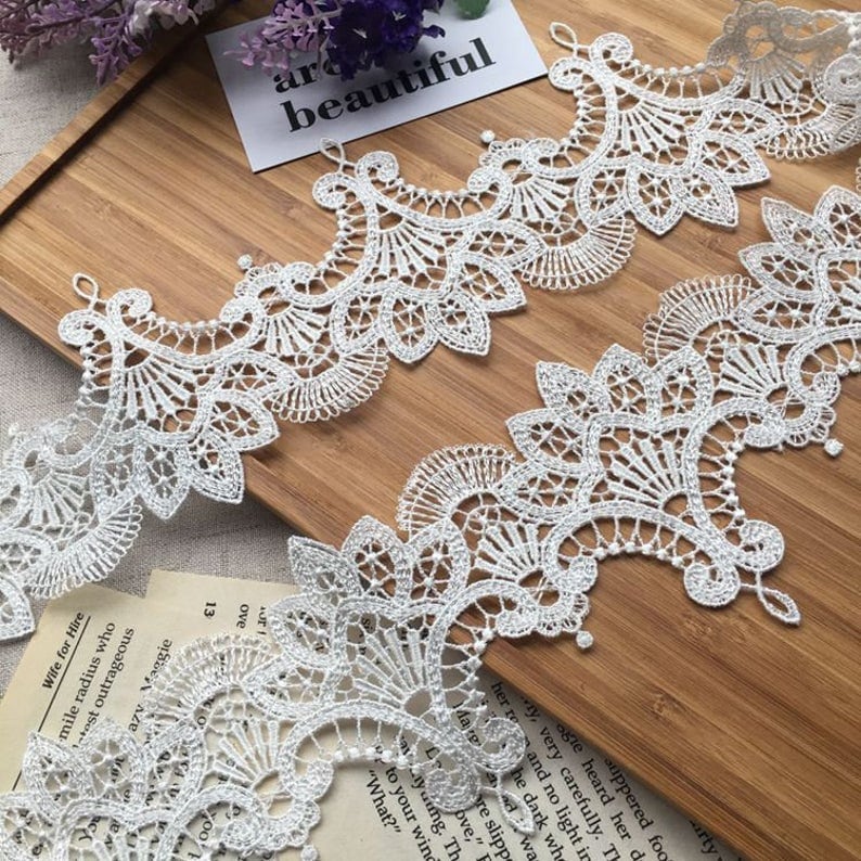 May include: Two pieces of white lace trim with a delicate floral pattern. The lace is arranged in a wave-like pattern on a wooden surface.