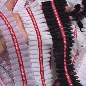 Puede incluir: Primer plano de ribete de tela con volantes en blanco y negro. Cada ribete presenta una cinta roja con un estampado floral. La tela está fruncida y en capas, creando un efecto texturizado. Ideal para proyectos de costura y manualidades.