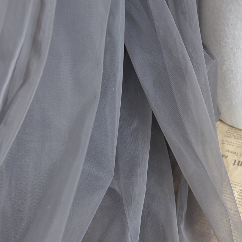 Pu&ograve; includere: Tessuto di tulle grigio con una texture morbida e trasparente. Il tessuto &egrave; piegato e drappeggiato, mostrando la sua qualit&agrave; delicata e ariosa.