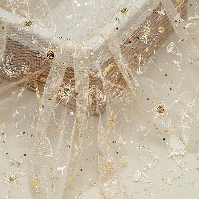 May include: Delicate ivory tulle fabric with gold embroidered celestial designs, including stars, suns, and floral accents. The fabric is embellished with scattered sequins, adding a subtle shimmer. Ideal for bridal wear, evening gowns, or decorative projects.