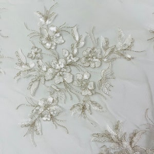 Könnte beinhalten: Weißer Blumenspitzenstoff mit silbernen Pailletten. Die Spitze hat ein zartes Blumenmuster mit kleinen, schimmernden Pailletten.