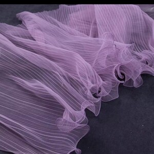 Peut inclure: Gros plan sur un tissu lavande fluide avec un design pliss&eacute;. Le tissu a une qualit&eacute; d&eacute;licate et transparente, avec de subtiles lignes verticales ajoutant de la texture. Les bords sont finis avec des d&eacute;tails fronc&eacute;s, cr&eacute;ant un aspect doux et &eacute;th&eacute;r&eacute;.
