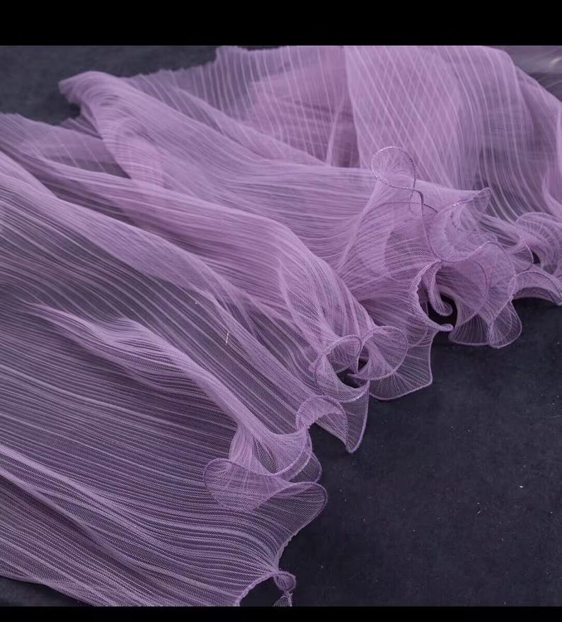 Peut inclure: Gros plan sur un tissu lavande fluide avec un design pliss&eacute;. Le tissu a une qualit&eacute; d&eacute;licate et transparente, avec de subtiles lignes verticales ajoutant de la texture. Les bords sont finis avec des d&eacute;tails fronc&eacute;s, cr&eacute;ant un aspect doux et &eacute;th&eacute;r&eacute;.