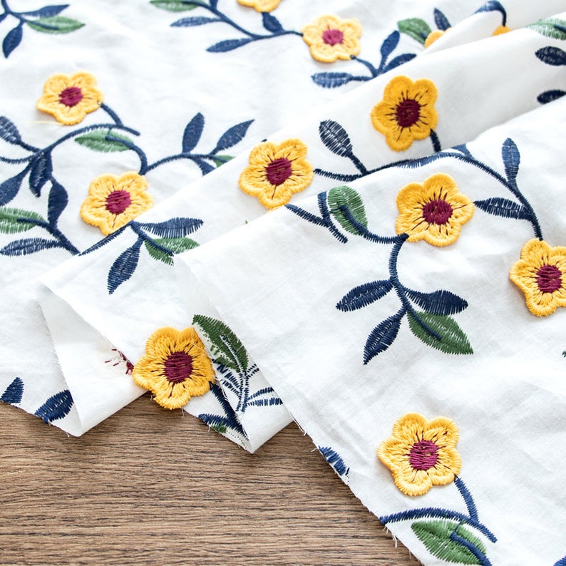 May include: White fabric with embroidered yellow flowers and green leaves. The fabric is folded and the flowers are in focus.