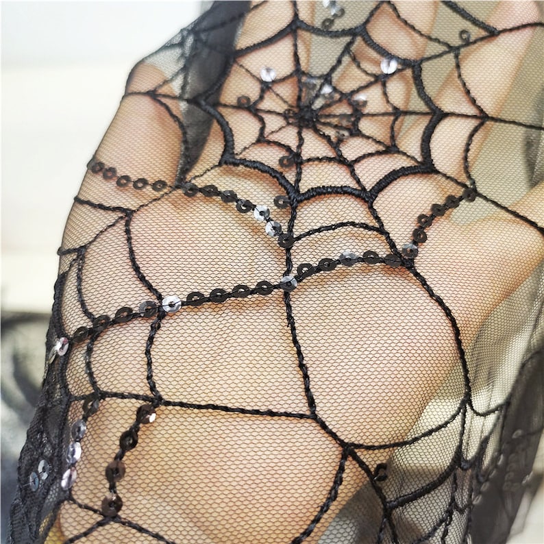 May include: A sheer black mesh fabric featuring an embroidered spider web pattern. The web design is accented with small, reflective silver sequins stitched along the lines of the web, creating a shimmering effect against the dark netting.