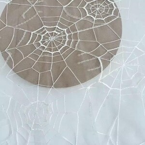 May include: A sheer white fabric featuring an intricate embroidered pattern of multiple spider webs. The delicate white thread creates detailed web designs across the translucent material, which is layered over a solid tan circular base.
