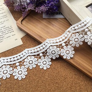 May include: White lace trim with a scalloped edge and a repeating pattern of small flowers and circles. The trim is shown on a wooden surface.