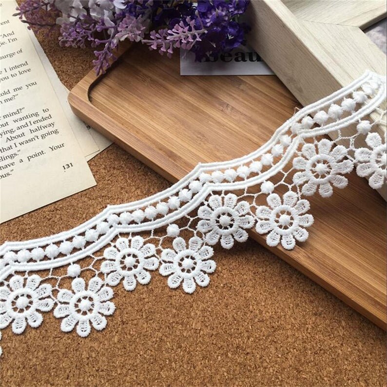 May include: White lace trim with a scalloped edge and a repeating pattern of small flowers and circles. The trim is shown on a wooden surface.