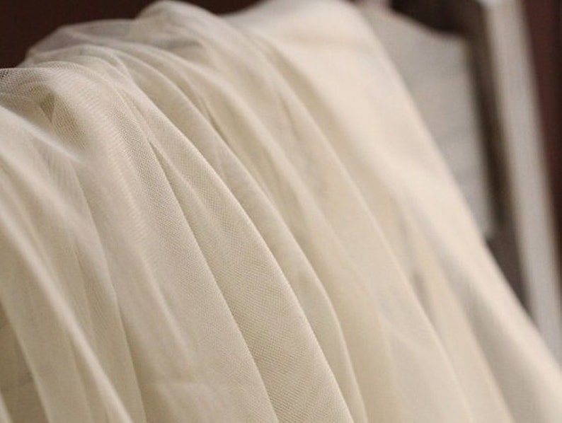 May include: Close-up of a white sheer fabric with a soft, flowing texture. The fabric is draped and folded, creating a delicate and airy appearance.