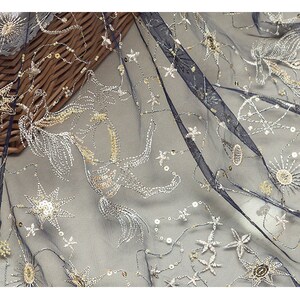 May include: Navy blue sheer fabric with embroidered celestial designs. The fabric features silver and gold star and sunburst patterns, with sequin accents. The embroidery creates a shimmering effect, suitable for apparel or decorative projects.