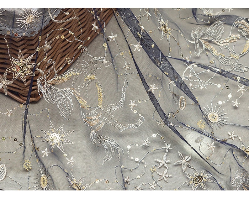 May include: Navy blue sheer fabric with embroidered celestial designs. The fabric features silver and gold star and sunburst patterns, with sequin accents. The embroidery creates a shimmering effect, suitable for apparel or decorative projects.