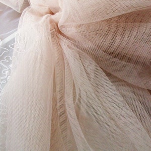 May include: A close-up of a pale pink tulle fabric with a fine mesh pattern. The fabric is soft and delicate, with a slightly shimmery finish.