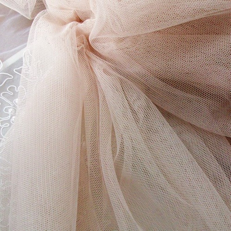 May include: A close-up of a pale pink tulle fabric with a fine mesh pattern. The fabric is soft and delicate, with a slightly shimmery finish.