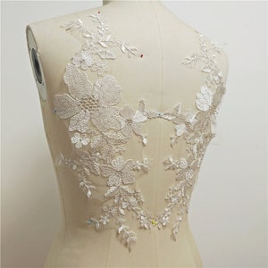 May include: White floral lace applique with pearl embellishments. The lace is delicate and intricate, with a floral design. The applique is perfect for adding a touch of elegance to any garment.