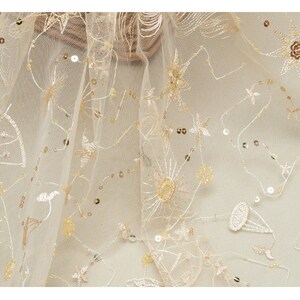 May include: Ivory-colored tulle fabric with embroidered celestial designs, including stars, suns, and other shapes. The fabric is embellished with sequins, adding a subtle shimmer. The embroidery and sequins are in shades of gold and beige.