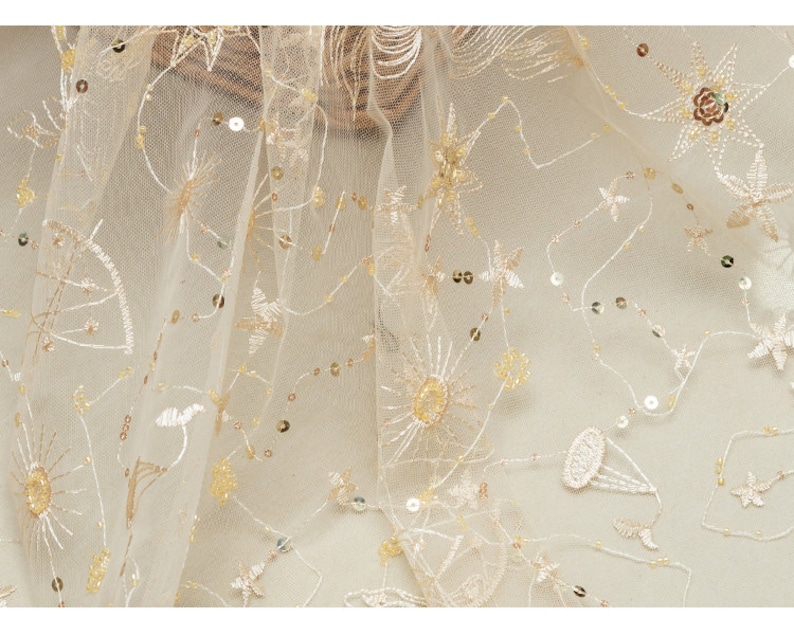 May include: Ivory-colored tulle fabric with embroidered celestial designs, including stars, suns, and other shapes. The fabric is embellished with sequins, adding a subtle shimmer. The embroidery and sequins are in shades of gold and beige.
