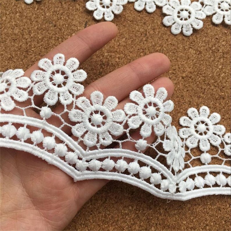 May include: White lace trim with a scalloped edge and a repeating floral pattern. The lace is delicate and intricate, with small flowers and a delicate net-like design.