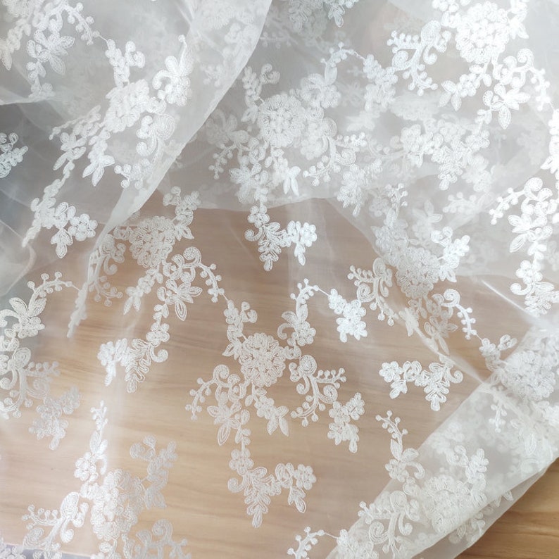 May include: White floral lace fabric with a delicate pattern. The lace is sheer and has a soft, flowing texture.