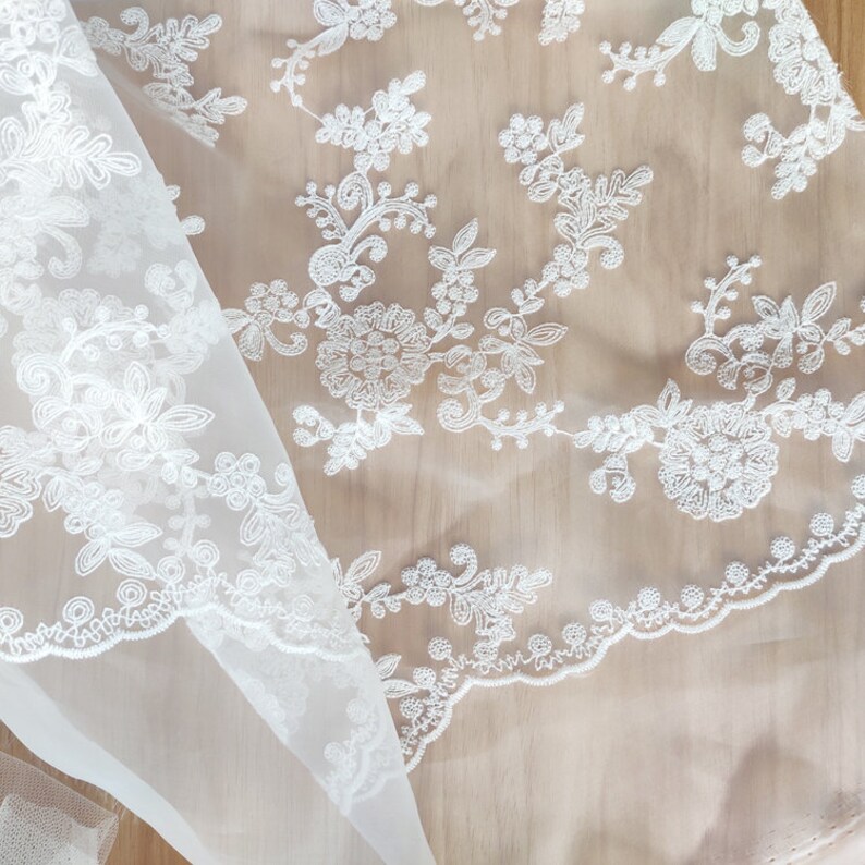 May include: White sheer fabric with delicate floral embroidery. The fabric is folded in half, showing the intricate details of the design.
