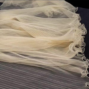 Peut inclure: Un tissu fluide de couleur cr&egrave;me avec une texture d&eacute;licate et transparente. Le mat&eacute;riau est fronc&eacute; et volants sur un bord, cr&eacute;ant un effet doux en forme de vague. Le tissu semble l&eacute;ger et a&eacute;r&eacute;, adapt&eacute; aux mariages ou aux tenues de soir&eacute;e.