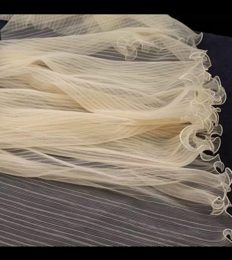 Peut inclure: Un tissu fluide de couleur cr&egrave;me avec une texture d&eacute;licate et transparente. Le mat&eacute;riau est fronc&eacute; et volants sur un bord, cr&eacute;ant un effet doux en forme de vague. Le tissu semble l&eacute;ger et a&eacute;r&eacute;, adapt&eacute; aux mariages ou aux tenues de soir&eacute;e.