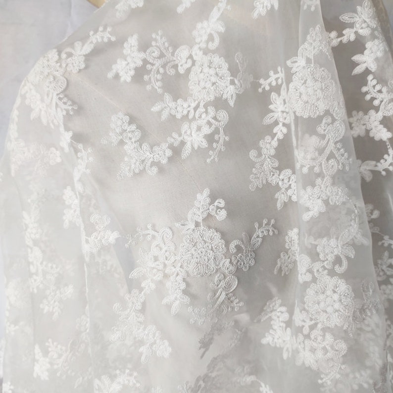 May include: White sheer fabric with delicate floral embroidery. The fabric is lightweight and translucent, with a soft, flowing texture.
