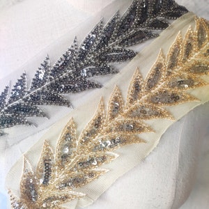 May include: Two rows of sequined trim, one gold and one silver, on a white fabric background. The trim features a repeating leaf design.