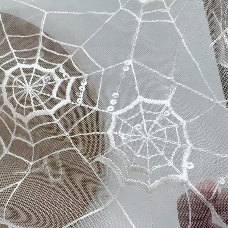 May include: A white sheer mesh fabric featuring an embroidered spider web pattern. The design includes delicate white thread stitching forming the webs, accented with small, clear, reflective sequins integrated along the lines of the web pattern.