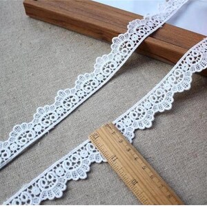May include: White lace trim with a scalloped edge. The lace is delicate and intricate, with a repeating pattern of small circles and loops. The trim is shown on a wooden surface with a ruler for scale.