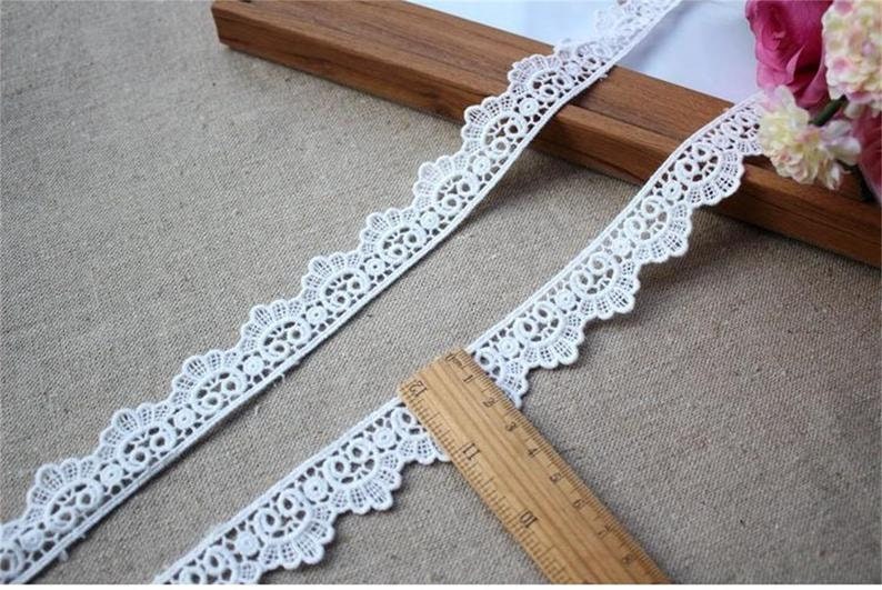 May include: White lace trim with a scalloped edge. The lace is delicate and intricate, with a repeating pattern of small circles and loops. The trim is shown on a wooden surface with a ruler for scale.