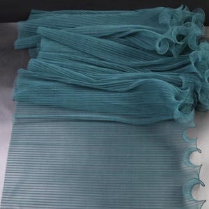 Peut inclure: Un empilement de tissu pliss&eacute; de couleur sarcelle avec un bord ondul&eacute; et festonn&eacute;. Le tissu a une texture c&ocirc;tel&eacute;e et est superpos&eacute;, cr&eacute;ant de la profondeur. La couleur est un sarcelle sourd, et le tissu semble l&eacute;ger et fluide.