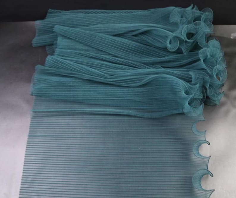 Peut inclure: Un empilement de tissu pliss&eacute; de couleur sarcelle avec un bord ondul&eacute; et festonn&eacute;. Le tissu a une texture c&ocirc;tel&eacute;e et est superpos&eacute;, cr&eacute;ant de la profondeur. La couleur est un sarcelle sourd, et le tissu semble l&eacute;ger et fluide.